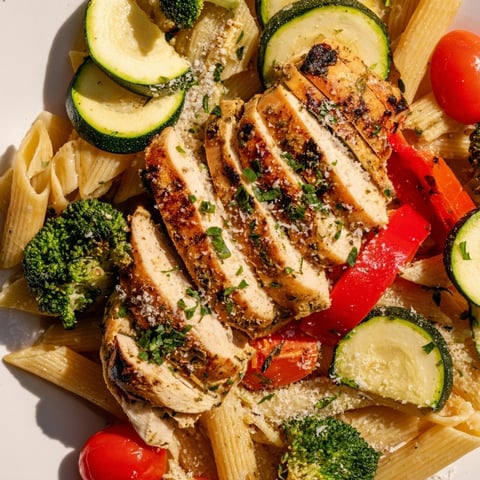 Golden-brown, juicy garlic herb chicken slices rest atop a vibrant skillet of penne, cherry tomatoes, zucchini, and broccoli in a light sauce.