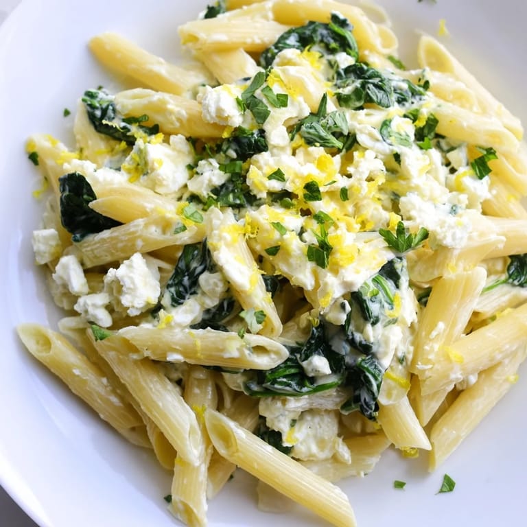Freshly grated lemon zest and chopped basil garnish this creamy feta spinach pasta, served warm on a rustic plate for family-style enjoyment.