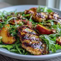 Grilled Chicken and Peach Arugula Salad with Balsamic Drizzle on a white plate, garnished with toasted pecans and feta cheese.